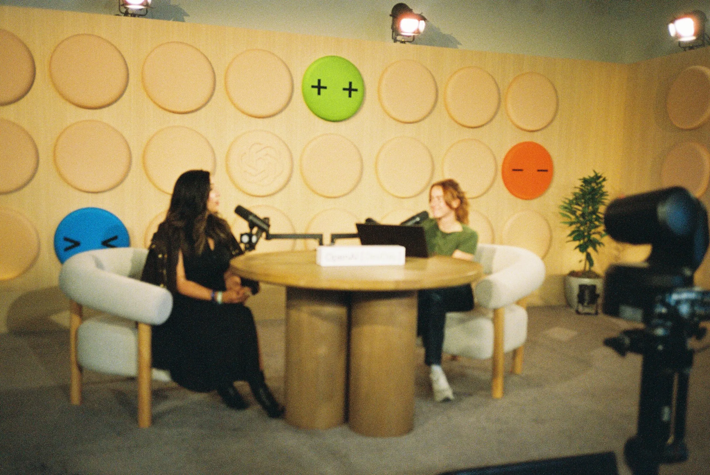 Two women in conversation on the podcast set, a camera rig in the foreground