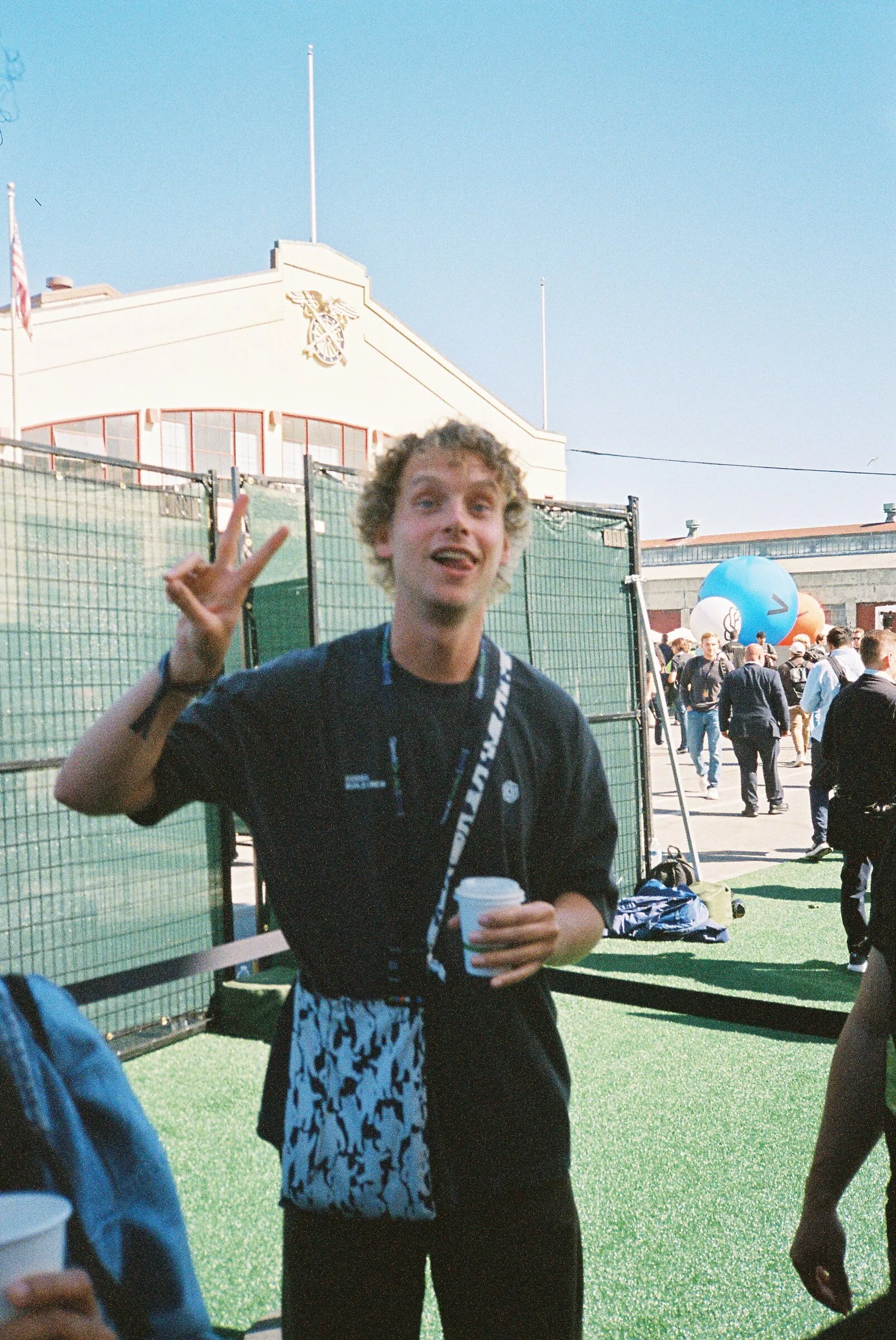A man wearing a lanyard flashing a peace sign outdoors, holding a cup