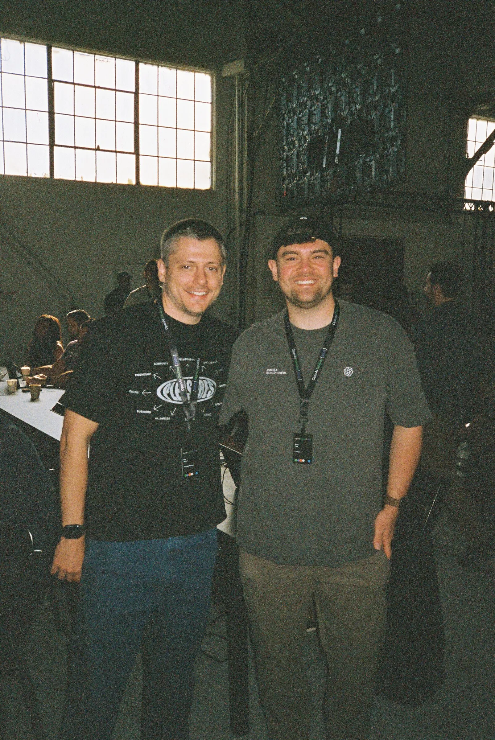 Two men standing together in a dim hall by tall windows