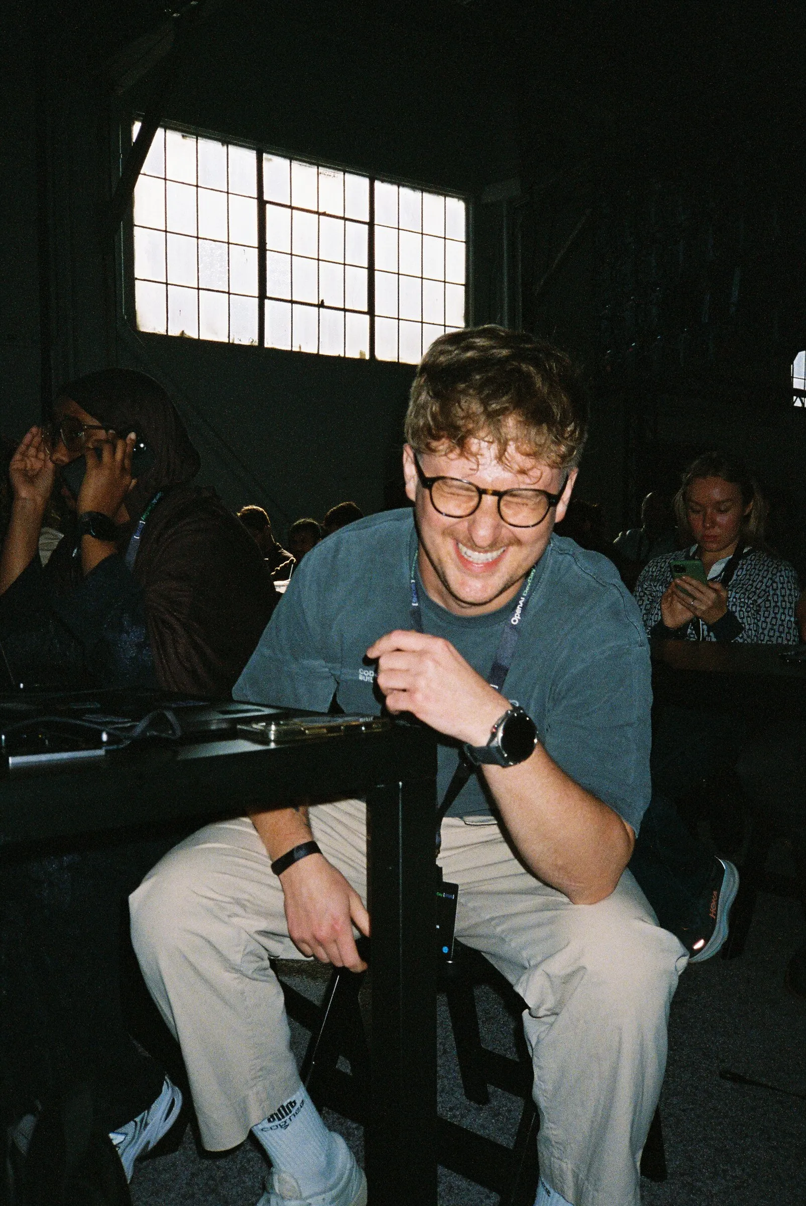 A man in glasses grinning, perched on a railing in a dark hall