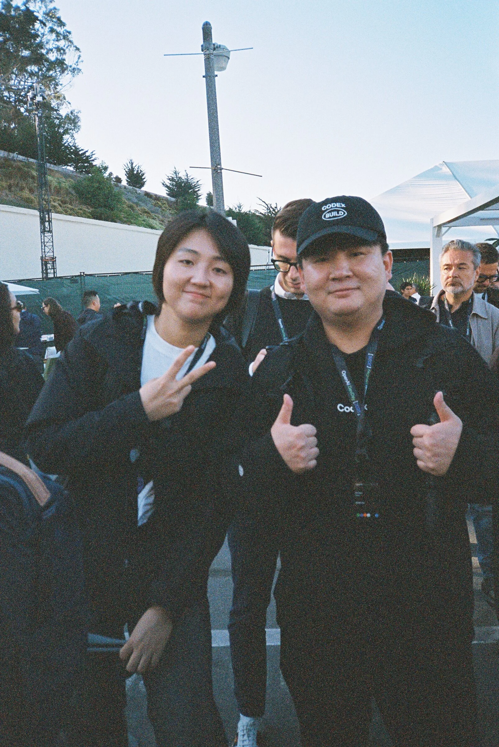 A woman and a man in a cap giving a peace sign and thumbs up outside