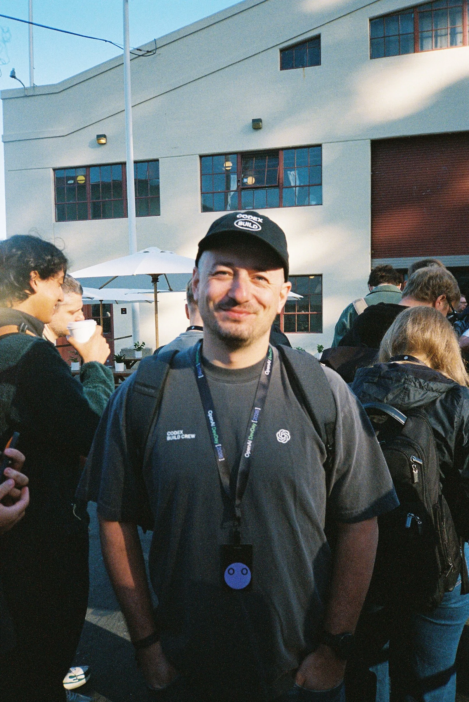 A man in a black cap and lanyard standing in a sunny courtyard