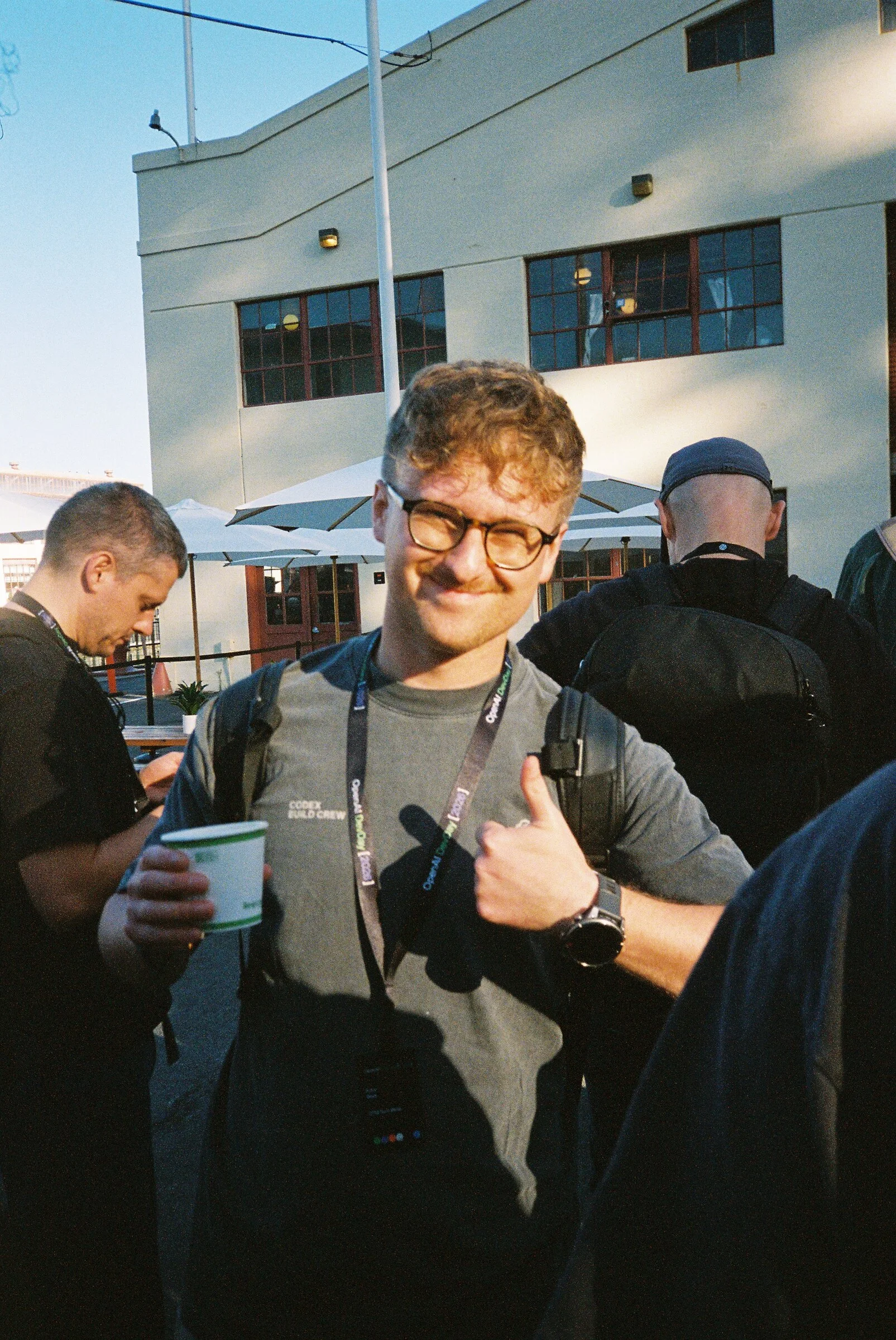 A man in glasses giving a thumbs up, holding a coffee