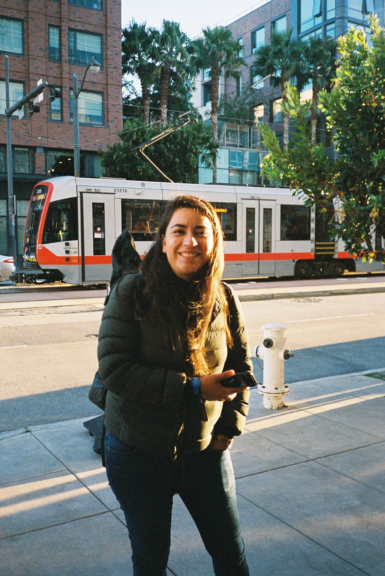A woman on a sunny pavement with a Muni train passing behind